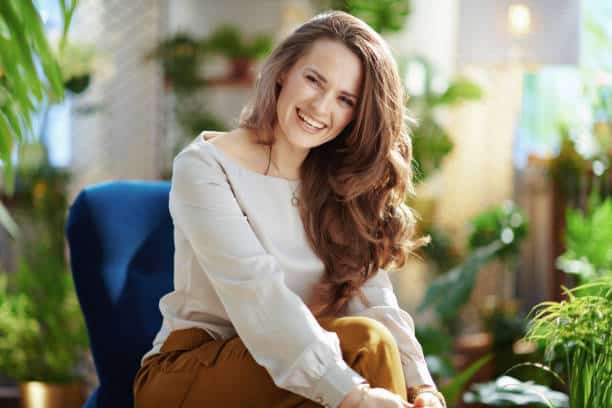 Green Home. Portrait of smiling trendy middle aged woman with long wavy hair in the modern house in sunny day in green pants and grey blouse sitting in a blue armchair. Happiness: The ultimate birthright