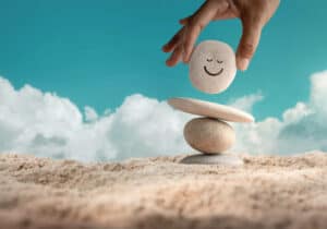 Enjoying Life Concept. Harmony and Positive Mind. Hand Setting Natural Pebble Stone with Smiling Face Cartoon to Balance on Beach Sand. Balancing Body, Mind, Soul and Spirit. Mental Health Practice. The Feeling of Emotions and Personal Growth