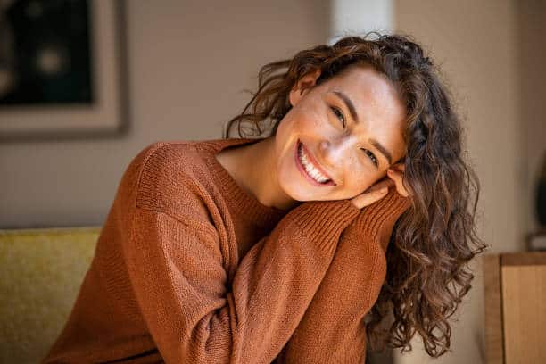 Happy young woman sitting on sofa at home and looking at camera. Portrait of comfortable woman in winter clothes relaxing on armchair. Portrait of beautiful girl smiling and relaxing during autumn. The five options to build happiness