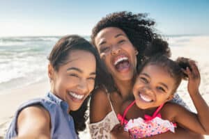 Portrait of smiling young african american woman with child taking selfie at beach with her best friend. Cheerful multiethnic gay couple enjoying at beach with daughter during summer holiday. Happy smiling young mixed race sisters with cute little girl taking selfie over exotic tropical beach. Laughter, play, fun, joy, and happiness