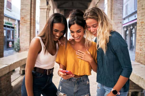Three happy women using mobile phone outdoors. Group of smiling female friends watching social media at smartphone. High quality photo. Laughter, play, fun, joy, and happiness
