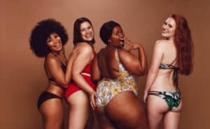Group of women of different race, figure and size in swimsuits standing together and laughing against grey background. Diverse women in bikinis looking at camera. Love Your Body, Forgive Your 