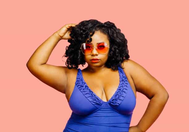 Portrait of a beautiful plus size model in a bathing suit and sunglasses, isolated on pink. Learning to love your curves