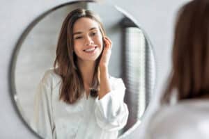 Beauty Concept. Portrait Of Attractive Happy Woman Looking At Mirror In Bathroom, Beautiful Millennial Lady Wearing White Silk Robe Smiling To Reflection, Enjoying Her Appearance, Selective Focus. Be happy happiness is just a matter of mind