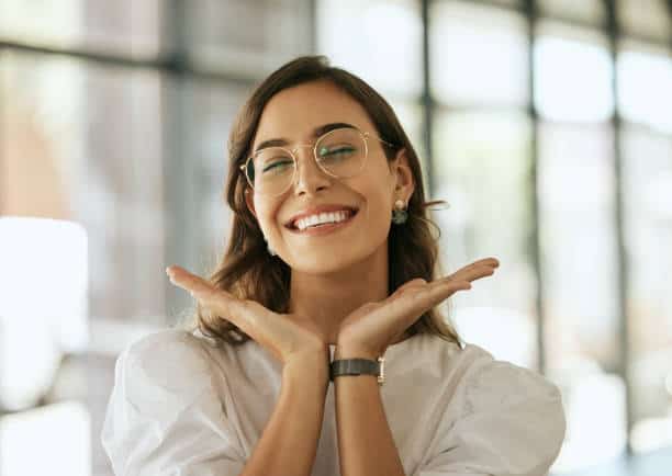 Cheerful business woman with glasses posing with her hands under her face showing her smile in an office. Playful hispanic female entrepreneur looking happy and excited at workplace. Going for Happiness