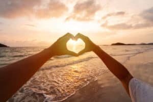 Young couple traveler making heart shape with hands on the beach at sunset, Lovers on honeymoon and Valentine's day concept. The heart of happiness. 