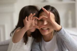 Happy young mother with cute little daughter making focused heart sign with hands, looking at camera. Smiling millennial mom and small girl showing love gesture together, expressing care, affection. The heart of happiness. 