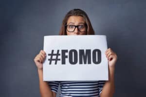 Studio shot of an attractive young woman holding a sign with #FOMO printed on it against a gray background. what are you really missing out on. 
