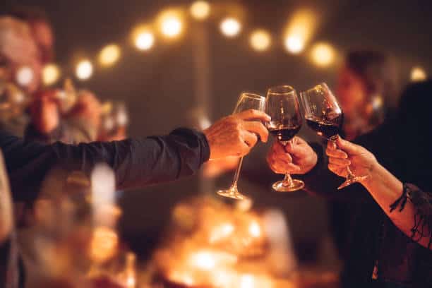 Celebratory red wine toast between senior adult friends at candle light social event party with string fairy lights