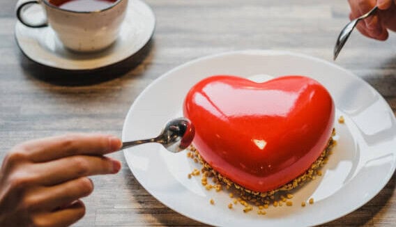 Two people sharing a heart-shaped red dessert on a white plate with spoons, accompanied by a cup of tea on a wooden table.